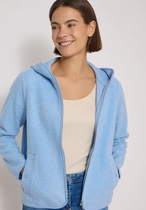 Lächelnde Frau in hellblauer Fleecejacke mit Kapuze, beigem Top und Jeans, Hände in den Taschen.
