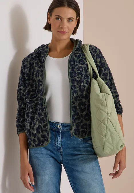 Femme en jean, débardeur blanc et veste à motif, portant sur l'épaule un grand sac matelassé vert.