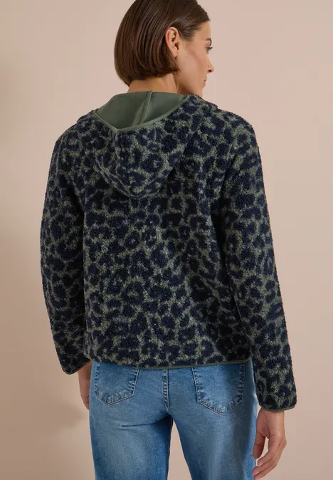 Vue de dos d’une femme portant un sweat à capuche en polaire bleu-vert à motifs et un jean clair sur fond beige.