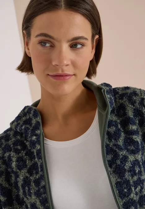 Une femme aux cheveux bruns courts porte une veste polaire verte à imprimé animalier et un t-shirt blanc.