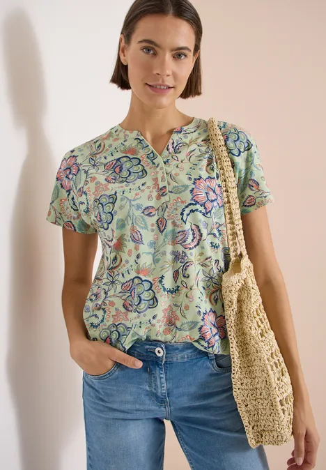 Shirt met split in de hals en print raw matcha lime Vrouw draagt gebloesd T-shirt, blauwe jeans en een strooien tas.