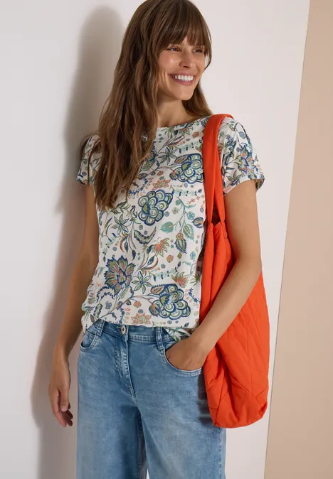 Frau lächelt, trägt gemustertes T-Shirt, Jeans und eine orangefarbene Umhängetasche.