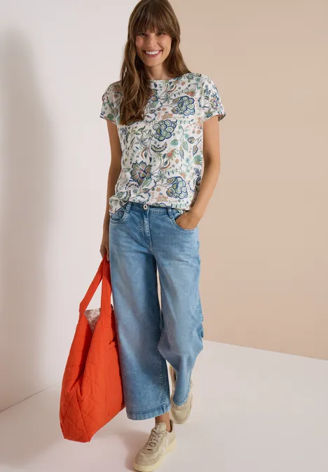 Frau mit geblümtem T-Shirt, weiten Jeans, beigen Sneakern und großer oranger Tasche.