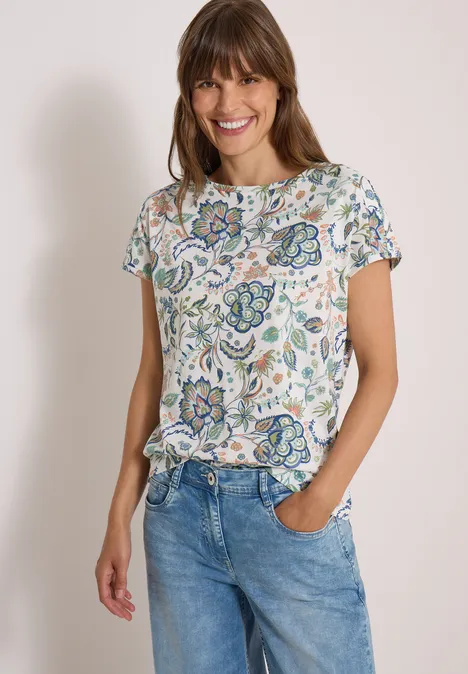 Eine lächelnde Frau trägt eine bedruckte Blumenbluse und blaue Jeans.