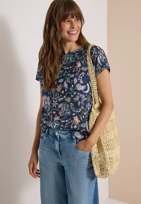 Lächelnde Frau im geblümten Shirt, Jeans und gehäkelter Tasche.