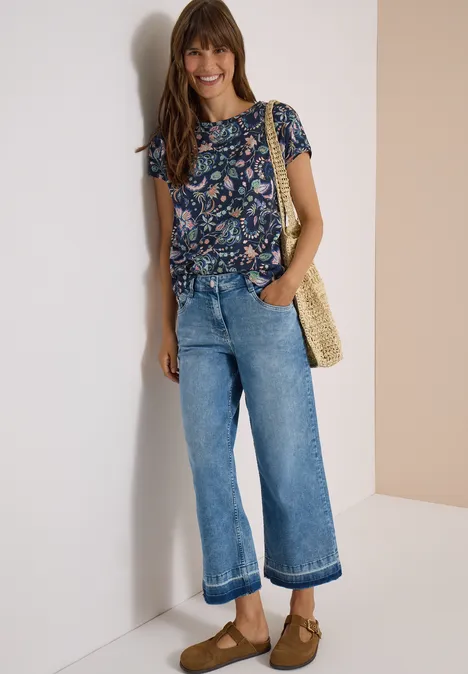 Frau in Blüten-Print-Bluse, weiten Jeans-Culotte, Strohtasche und braunen Clogs, lächelnd an weißer Wand.