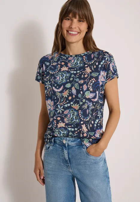 Lächelnde Frau mit blauem Shirt im Blumenprint und hellen Jeans, eine Hand in Tasche.