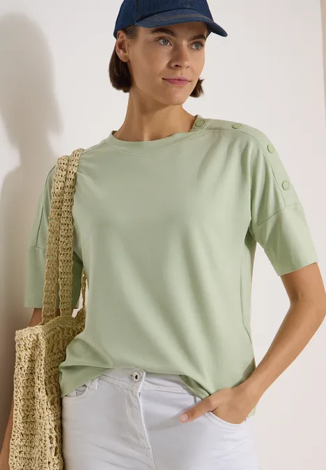 Dropped Shoulder Shirt mit Knöpfen raw matcha lime Frau in mintgrünem T-Shirt, weißer Hose, Strohtasche und Cap vor heller Wand.
