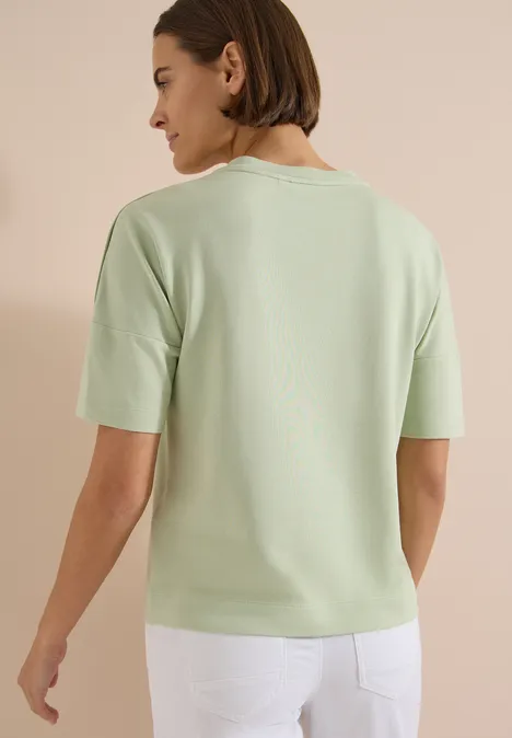 Dropped Shoulder Shirt mit Knöpfen raw matcha lime Frau von hinten in hellgrünem Kurzarmoberteil und weißer Hose.