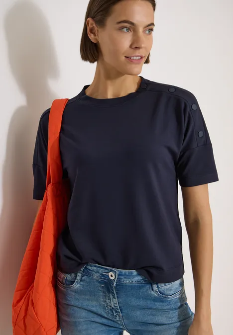 Chemise à épaules tombantes avec boutons urban dark blue Frau in dunkelblauem T-Shirt mit Schulterknöpfen, Jeans und roter Tasche vor heller Wand.