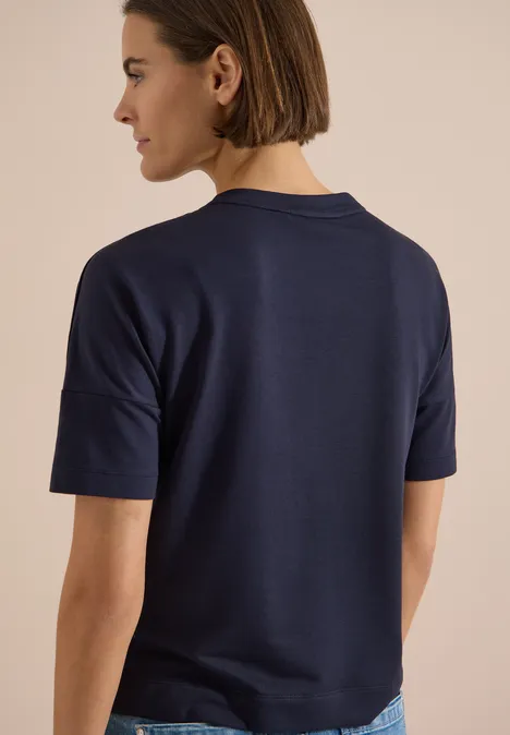 Chemise à épaules tombantes avec boutons urban dark blue Rückansicht einer Frau in navyblauem T-Shirt und Jeans vor hellem Hintergrund.