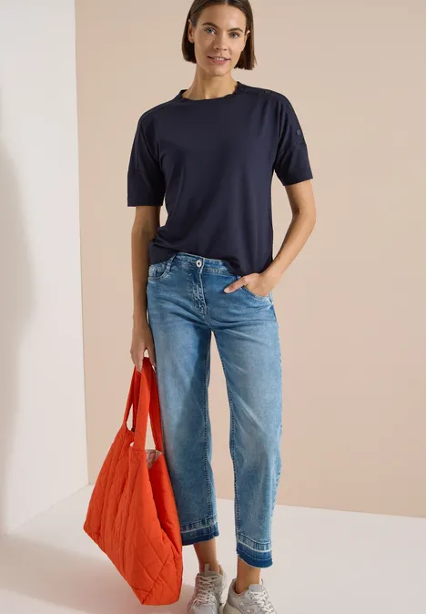 Chemise à épaules tombantes avec boutons urban dark blue Person in dunkelblauem T-Shirt, hellblauen Jeans und oranger Stepp-Tasche vor beigefarbenem Hintergrund.