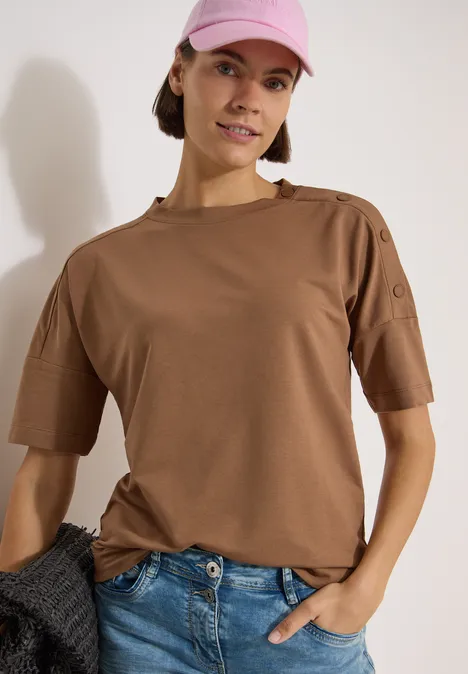Dropped Shoulder Shirt mit Knöpfen creamy mocha Person in braunem Kurzarmshirt mit Zierknöpfen auf der Schulter, blaue Jeans, eine Hand in der Hosentasche.