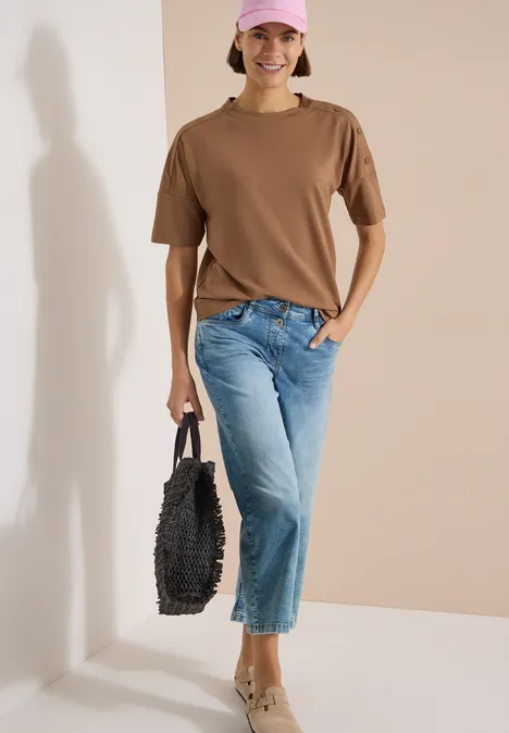 Dropped Shoulder Shirt mit Knöpfen creamy mocha Person in braunem T-Shirt, blauen Jeans und schwarzer Tasche vor hellem Hintergrund.