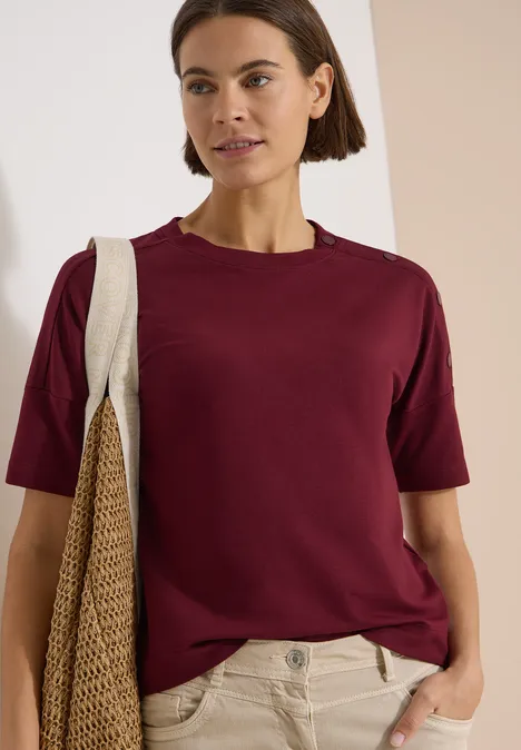 Dropped Shoulder Shirt mit Knöpfen red wine Frau trägt weinrotes T-Shirt, beige Hose und beigen Schulterbeutel vor neutralem Hintergrund.