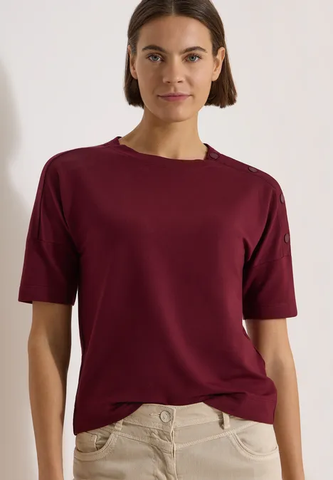 Dropped Shoulder Shirt mit Knöpfen red wine Eine Frau in dunkelrotem T-Shirt mit kurzen Ärmeln und heller Hose vor neutralem Hintergrund.