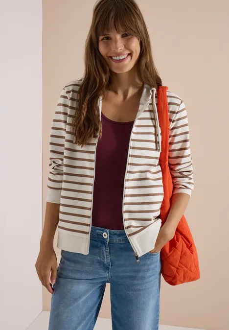 Frau in gestreifter Jacke, burgunderrotem Top, Jeans und oranger Tasche lächelt