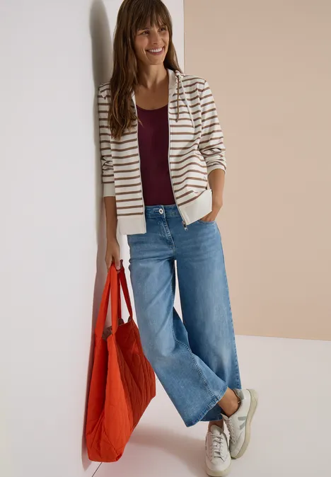 Frau in gestreiftem Blazer, Bordeauxshirt, weitem Jeans, Sneakers, trägt roten Stoffbeutel.