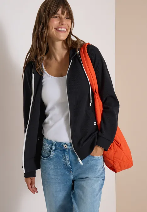 Eine lächelnde Frau in weißem Top, schwarzer Kapuzenjacke, Jeans und orangener Tasche.