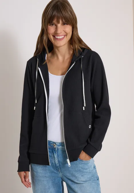 Eine lächelnde Frau trägt eine schwarze Kapuzenjacke über weißem T-Shirt und blauen Jeans vor neutralem Hintergrund.