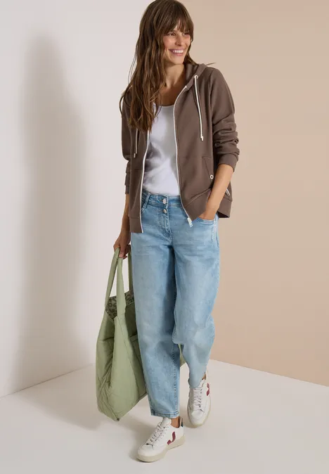 Frau im lässigen Look mit brauner Kapuzenjacke, weißem Shirt, hellblauen Jeans, weißen Sneakern und grüner Tasche.