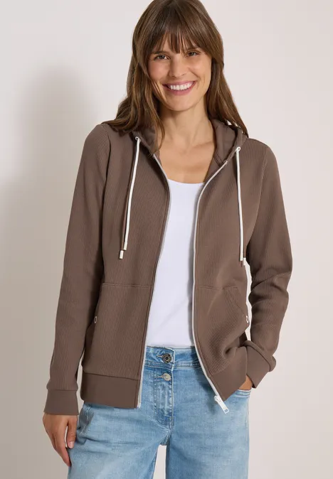 Eine Frau in brauner Kapuzenjacke, weißem Top und Jeans lächelt entspannt vor neutralem Hintergrund.