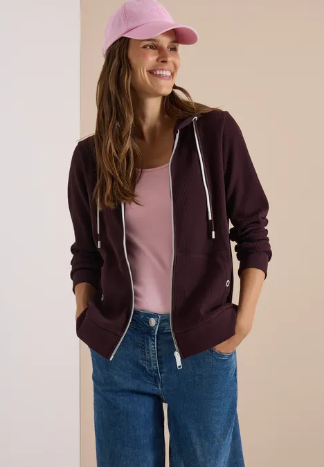 Veste à manches longues, à capuche et texturée berry red Eine lächelnde Frau in rosa Basecap, dunkelroter Kapuzenjacke, rosa Top und blauen Jeans vor neutralem Hintergrund.
