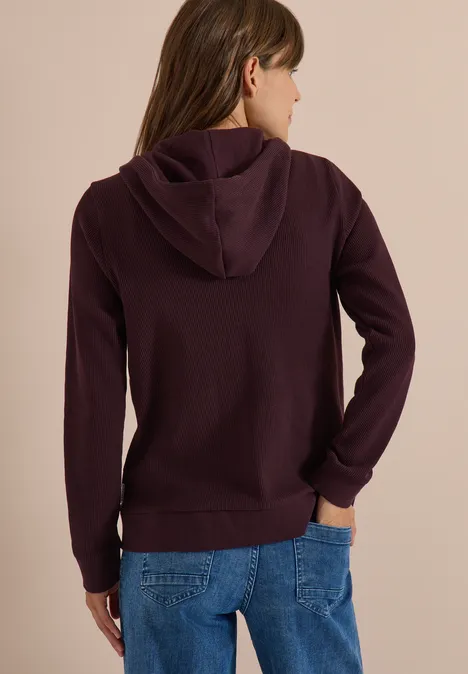 Veste à manches longues, à capuche et texturée berry red Person von hinten, trägt dunkelvioletten Kapuzenpullover und blaue Jeans.