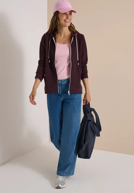Veste à manches longues, à capuche et texturée berry red Frau in pinkem Top, violetter Kapuzenjacke, weiter Jeans und Silber-Sneakern, rosafarbener Cap, sie hält eine blaue Jacke.