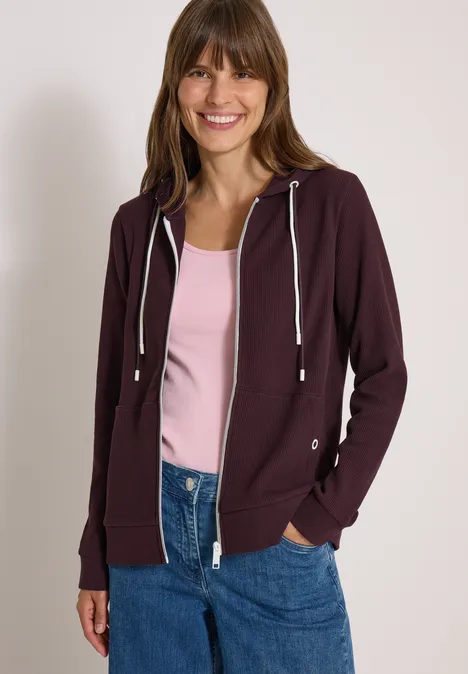 Veste à manches longues, à capuche et texturée berry red Junge Frau lächelt in einem dunkelvioletten Hoodie, rosa Top und Jeans vor weißer Wand.