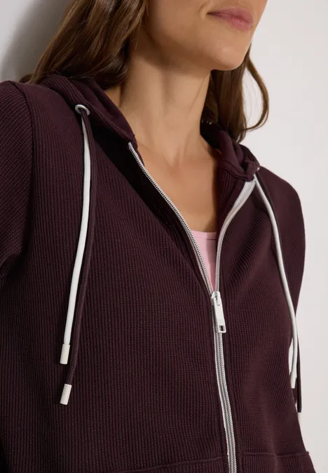 Veste à manches longues, à capuche et texturée berry red Frau trägt plumfarbenen Kapuzenpullover mit Reißverschluss und weißen Kordeln über hellem Shirt.