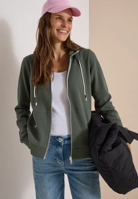 Veste à manches longues, à capuche et texturée dried olive green Frau trägt grünen Kapuzenpullover, weiße Weste, Jeans und rosa Cap, hält schwarze Jacke, lächelt.