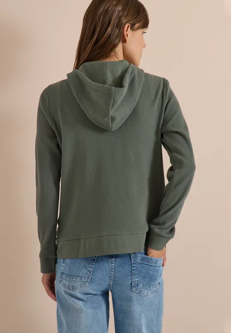 Veste à manches longues, à capuche et texturée dried olive green Person von hinten in grüner Kapuzenjacke mit Jeans.