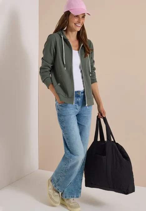 Veste à manches longues, à capuche et texturée dried olive green Junge Frau in Jeans, Hoodie und rosa Cap, die eine große schwarze Tasche trägt.
