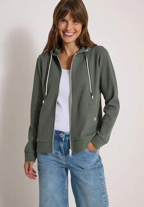 Veste à manches longues, à capuche et texturée dried olive green Lächelnde Frau in grüner Kapuzenjacke und Jeans vor hellem Hintergrund.