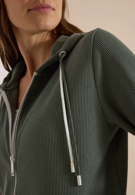 Veste à manches longues, à capuche et texturée dried olive green Nahaufnahme einer Person in einem olivgrünen, feinstrukturigen Hoodie mit Reißverschluss und hellem Kordelzug.