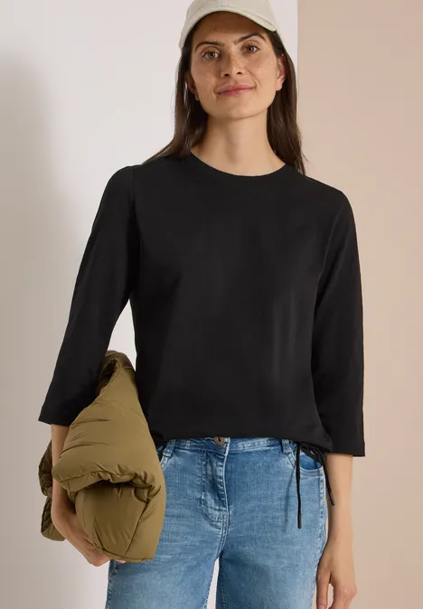3/4-Arm Shirt mit Rundhals in Unifarbe Black Frau in schwarzem Langarmshirt, Jeans und Baseballcap trägt eine grüne Steppjacke über dem Arm vor neutralem Hintergrund.