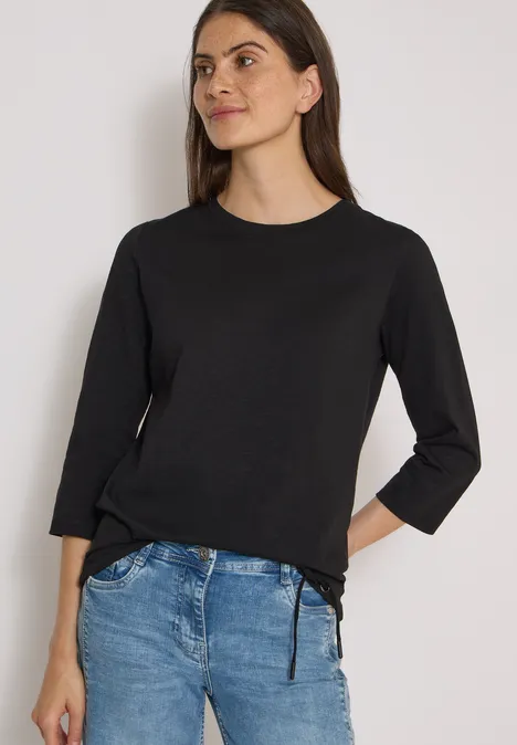 3/4-Arm Shirt mit Rundhals in Unifarbe Black Eine Frau in schwarzem 3/4-Arm-Shirt und blauen Jeans steht locker vor weißem Hintergrund.