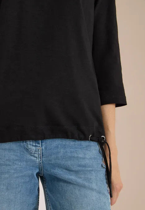 3/4-Arm Shirt mit Rundhals in Unifarbe Black Person trägt blaue Jeans und schwarzen Dreiviertelärmel-Pullover mit seitlicher Kordel, vor beigem Hintergrund.