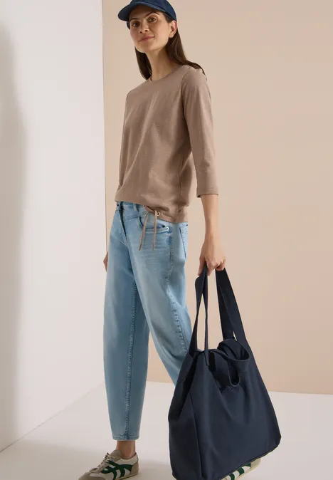 3/4-Arm Shirt mit Rundhals in Unifarbe natural taupe Frau in lässigem Outfit mit beiger Bluse, hellblauen Jeans und Cap hält eine große dunkelblaue Tasche.