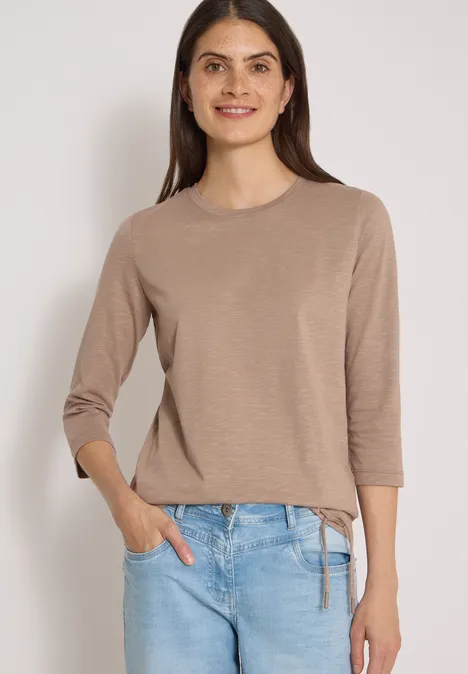 3/4-Arm Shirt mit Rundhals in Unifarbe natural taupe Frau lächelt in beigem 3/4-Arm-Pullover mit Seitenbindung und blauen Jeans vor hellem Hintergrund.