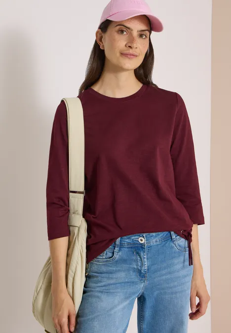 3/4-Arm Shirt mit Rundhals in Unifarbe red wine Junge Frau in weinrotem Dreiviertelarm-Shirt, blauen Jeans, rosa Cap und beiger Umhängetasche vor weißem Hintergrund.