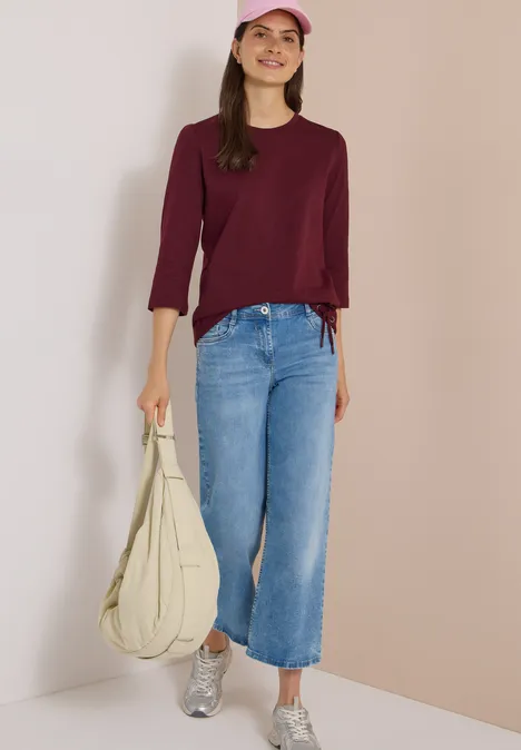 3/4-Arm Shirt mit Rundhals in Unifarbe red wine Frau in burgunderrotem Pullover, hellblauen Jeans, silbernen Sneakern, rosa Cap und beiger Tasche.