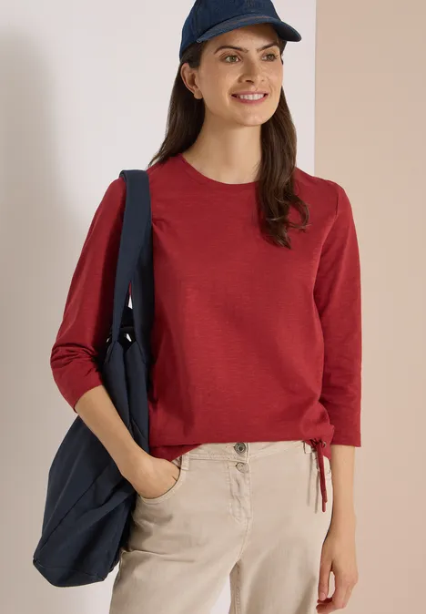 3/4-Arm Shirt mit Rundhals in Unifarbe russet red Junge Frau mit rotem Pullover, beiger Hose und blauem Cap trägt lächelnd eine blaue Tasche.