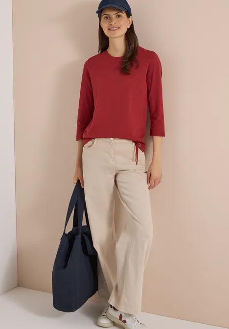 3/4-Arm Shirt mit Rundhals in Unifarbe russet red Person trägt rotes Oberteil, beige Hose, blaue Cap und hält blaue Tasche vor neutralem Hintergrund.