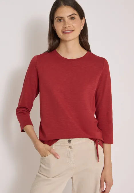 3/4-Arm Shirt mit Rundhals in Unifarbe russet red Frau trägt roten Dreiviertelarm-Pullover und helle Chinohose, steht lächelnd mit Händen in den Taschen.