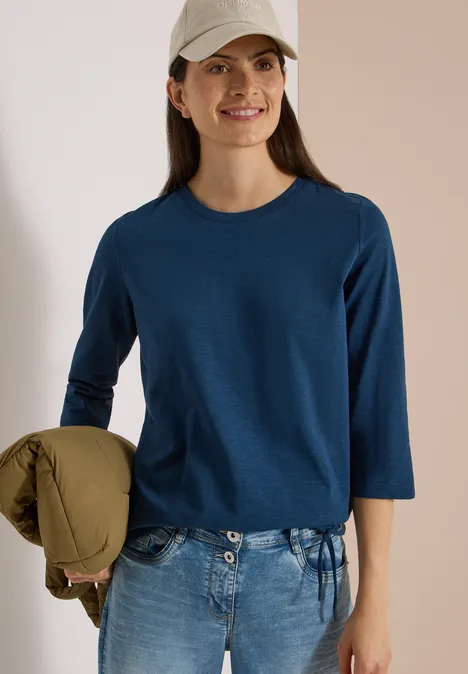 3/4-Arm Shirt mit Rundhals in Unifarbe moonlight blue Lächelnde Frau mit Cap, blauem Dreiviertelshirt und Jeans, hält locker eine Jacke.