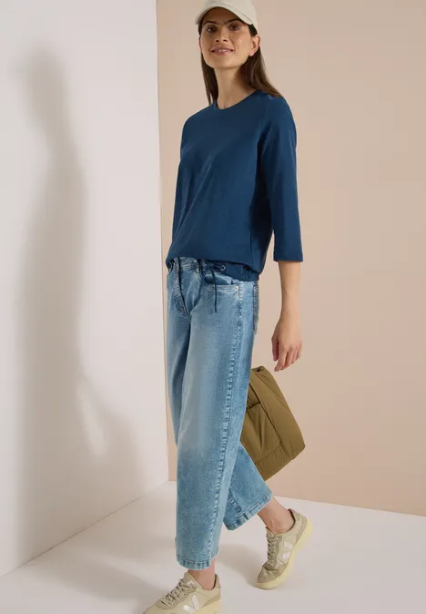 3/4-Arm Shirt mit Rundhals in Unifarbe moonlight blue Frau in blauem Shirt, weit geschnittenen Jeans, Sneakern und Cap, trägt eine grüne Tasche.
