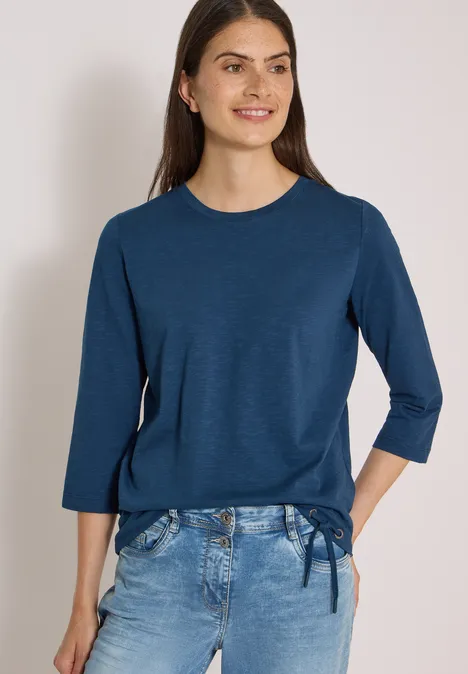 3/4-Arm Shirt mit Rundhals in Unifarbe moonlight blue Frau trägt marineblaues 3/4-Arm-Shirt mit seitlicher Schleife und helle Jeans, lächelnd vor hellem Hintergrund.