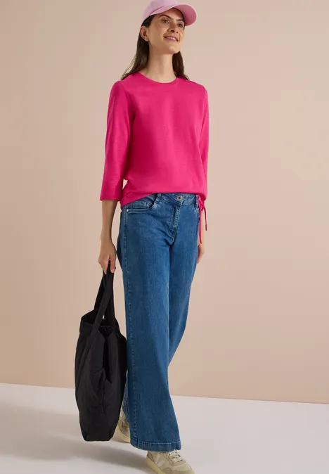 3/4-Arm Shirt mit Rundhals in Unifarbe pink flash Frau in pinkem Top, weiter blauer Jeans, pinker Cap und schwarzer Tasche vor neutralem Hintergrund.
